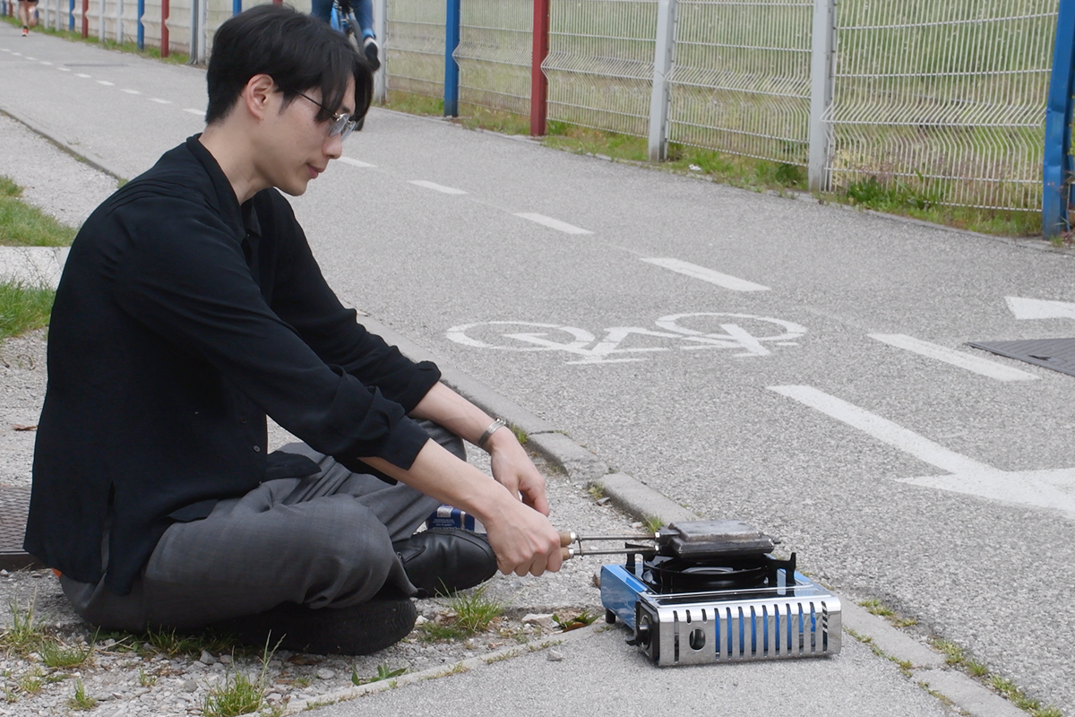 Rui Yamaguchi sitting by the road along the border, toasting a hot sandwich on a stove