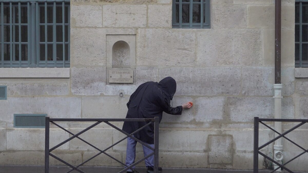 Demonstration for becoming a “professional unemployed” – the hooded artist wiping the wall beside the Louis Hélie memorial plaque with an orange cloth, Rue de Seine, Paris