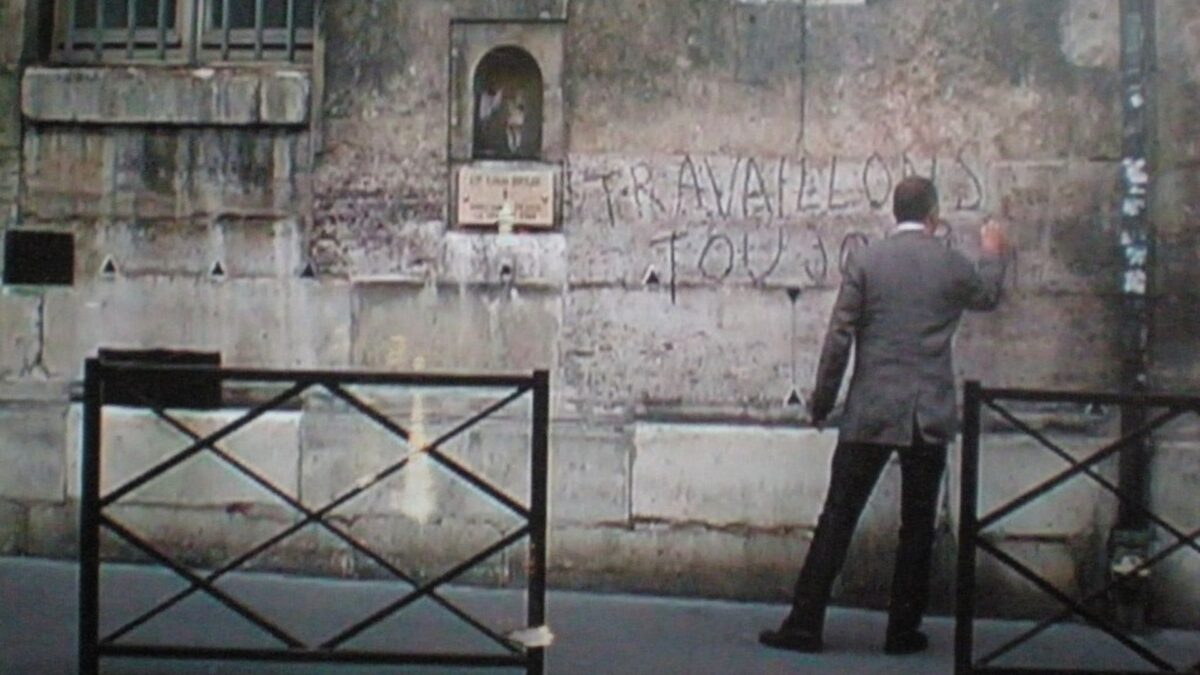 Video still from Marianne Heier’s Jamais-toujours – a man writing “TRAVAILLONS TOUJOURS” on the same wall
